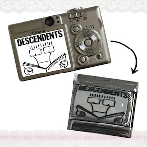 Könnte beinhalten: Eine silberne Canon Digitalkamera mit einem Aufkleber mit dem Wort "DESCENDENTS" und einer Comicfigur, die ein Buch liest. Der Aufkleber enthält auch den Satz "EVERYTHING SUCKS". Ein zweiter Aufkleber ist ebenfalls abgebildet.