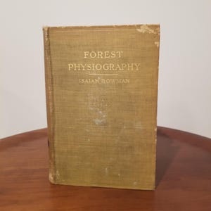 Puede incluir: Un libro antiguo titulado "FOREST PHYSIOGRAPHY" de Isaiah Bowman. La portada es de color verde oliva descolorido con letras doradas. El libro está sobre una superficie de madera.
