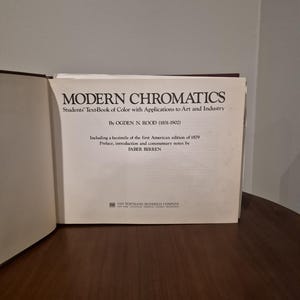 Puede incluir: Un libro abierto titulado "MODERN CHROMATICS" de Ogden N. Rood. La portada del libro es blanca con texto negro, incluyendo el subtítulo "Students' Text-Book of Color with Applications to Art and Industry". El libro incluye un facsímil de la primera edición americana de 1879.