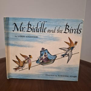 May include: Vintage children's book titled "Mr. Biddle and the Birds" by Lonzo Anderson, illustrated by Adrienne Adams. The cover features a whimsical illustration of a person in a boat being pulled by birds against a blue sky background.