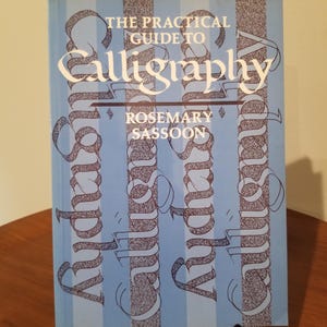 May include: A book titled "The Practical Guide to Calligraphy" by Rosemary Sassoon. The cover is light blue with vertical stripes and the word "Calligraphy" in large white script. The word "Calligraphy" is repeated vertically in a decorative font.
