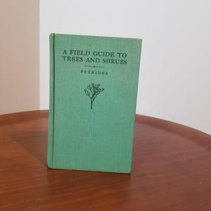 Könnte beinhalten: Ein grünes Hardcover-Buch mit dem Titel "A Field Guide to Trees and Shrubs" von Petrides. Das Buch hat eine Baumillustration und steht auf einer Holzoberfläche.