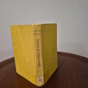 May include: A vintage book titled "Getting Organized" by Winston. The book has a yellow cover and spine with the title printed vertically. The book is standing on a dark brown surface.