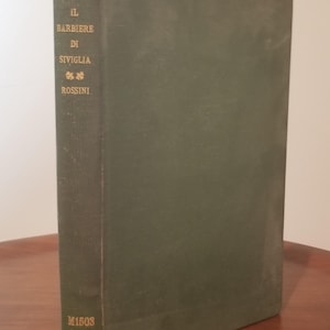 May include: A vintage, olive green book titled "Il Barbiere Di Siviglia" by Rossini. The spine features gold lettering and decorative elements. The book's bottom edge displays cataloging information and the year 1951.