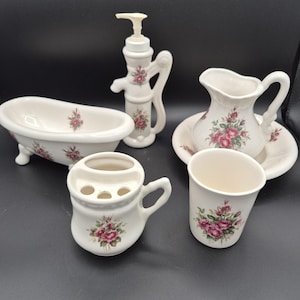 May include: A five-piece ceramic bathroom accessory set featuring a white base with pink rose floral patterns. The set includes a miniature clawfoot bathtub, a pump soap dispenser, a pitcher on a basin, a toothbrush holder, and a drinking cup.