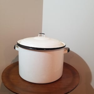 May include: White enamel pot with a lid, featuring black trim and handles. The pot is speckled and sits on a wooden side table with copper-colored legs. The pot is a kitchenware item.