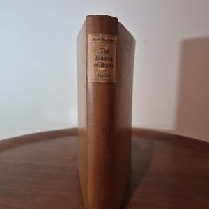 May include: A vintage book titled "The Binding of Books" by Horne. The book has a brown cover with a rectangular label on the spine. The label reads "Books about Books" at the top, with the title and author's name below. The book is standing upright on a wooden surface.