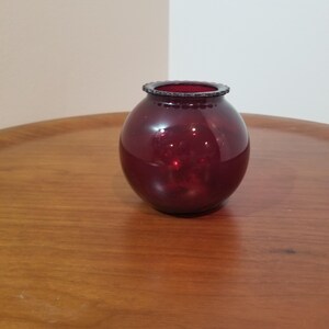 Peut inclure: Un petit vase rond en verre rouge rubis avec un bord texturé. Le vase est posé sur une surface en bois. Le vase a une forme sphérique et une couleur rouge foncé, adapté pour exposer de petites fleurs ou des objets décoratifs.