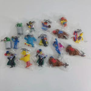 May include: A selection of Sesame Street character figurines in sealed plastic packaging. The figurines include characters like Big Bird, Elmo, and Cookie Monster, in various poses. The packaging has the words "Sesame Street" printed on it.
