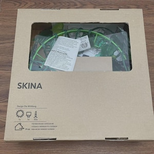 May include: A brown cardboard box with the name "SKINA" printed on it. The box has a window showing a green, wire-framed decoration with artificial leaves. Text in multiple languages, including "PRECAUCIÓN" and "FOR INDOOR AND OUTDOOR USE", is also visible.