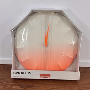 May include: A round wall clock with a white to orange gradient face and bright orange hands. The clock is in its original packaging, which has the word "SPRALLIS" printed on the front.