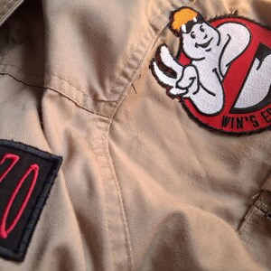 以下が含まれることがあります： ベージュ色のゴーストバスターズの制服のクローズアップ。制服には、ゴーストバスターズのロゴと「WIN'S ECTO VEGAS」の文字が入ったパッチが付いています。赤色の数字「220」が入った黒いパッチも表示されています。