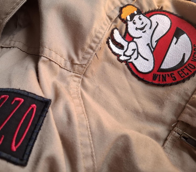 以下が含まれることがあります： ベージュ色のゴーストバスターズの制服のクローズアップ。制服には、ゴーストバスターズのロゴと「WIN'S ECTO VEGAS」の文字が入ったパッチが付いています。赤色の数字「220」が入った黒いパッチも表示されています。