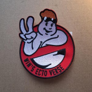 May include: Embroidered patch with a cartoon figure giving a peace sign, inside a red circle with a black and white smiling mouth. The text "WIN'S ECTO VERSE" is arched around the bottom of the circle.