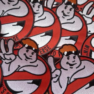 以下が含まれることがあります： オレンジ色の帽子と笑顔の顔をした漫画のキャラクターが描かれた刺繍パッチ。黒い縁取りのある赤い円が背景にあります。パッチには「VERSE」の文字が入っています。