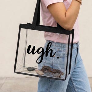Può includere: Borsa tote trasparente con bordi e manici neri, con la parola "ugh" in un carattere scritto a mano. La borsa contiene occhiali da sole, chiavi e un portachiavi.