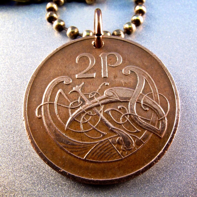 May include: A copper-colored 2 pence coin from the United Kingdom with a Celtic knot design featuring a bird. The coin is on a chain and can be used as a pendant.