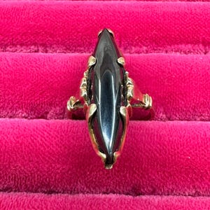 以下が含まれることがあります： 黒色のファセットカットのマルキーズ型の宝石がついた金の指輪。指輪は、細部までこだわった装飾的なデザインです。