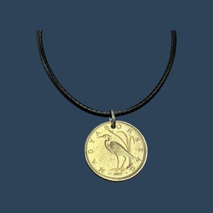 May include: A gold coin necklace with a black cord. The coin features a bird with a long neck and long legs standing on a branch. The coin has the text "GUYANA" and "1967" on it.