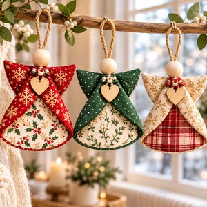May include: Three fabric angel ornaments hanging from a branch. Each features a wooden bead head, a jute rope loop, and a small wooden heart charm. The first is red with gold snowflakes, the second green with white dots, and the third cream with gold snowflakes and a red plaid base.
