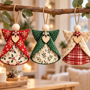 May include: Three fabric angel ornaments hanging from a branch. Each features a wooden bead head, a small wooden heart, and a jute twine loop. The left angel is red with gold snowflakes and holly. The center is green with white dots and foliage. The right is cream with gold snowflakes and a red plaid skirt.