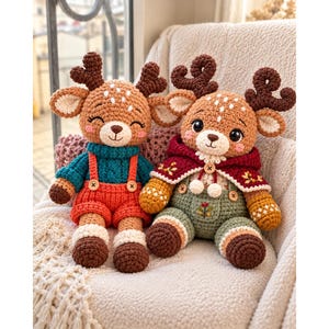 Pode incluir: Dois peluches de veado feitos em croché com hastes castanho-escuras e pelo castanho-claro. O veado da esquerda usa uma camisola azul-petróleo e jardineiras cor de laranja. O da direita usa uma capa com capuz bordeaux e jardineiras verdes.