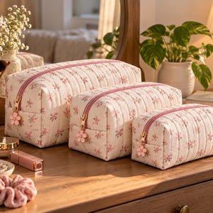 Könnte beinhalten: Ein Set aus drei rechteckigen, gesteppten Kosmetiktaschen in verschiedenen Größen aus cremefarbenem Stoff mit kleinem rosa Blumenmuster. Jede Tasche hat einen rosa Reißverschluss mit einem goldenen, blumenförmigen Anhänger.