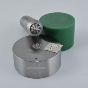 May include: A metal jewelry making tool set featuring a cylindrical stamp with a flower design, a round metal base, a green wax cylinder, and a small metal square with a stamped flower. The tools are for jewelry making.