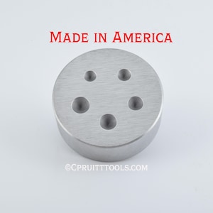 May include: A circular, silver-colored metal tool with five evenly spaced holes. The text "MADE IN AMERICA" is printed in red above the tool. The tool has a brushed metal finish and is likely used for jewelry making or metalworking.