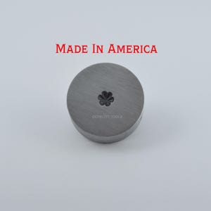 May include: A circular metal tool with a clover-shaped design in the center. The text "MADE IN AMERICA" is printed in red above the tool. The tool is made of a gray metal and has the text "©CPRUITT TOOLS" printed on it.