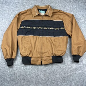 May include: A brown jacket with a black stripe across the chest, featuring an embroidered design. The jacket has a zip-up front, a collar, and black cuffs and waistband. The jacket is laid flat on a gray surface.