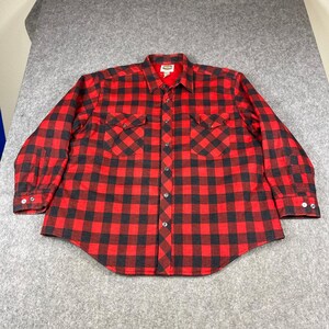 May include: A long-sleeve button-down shirt featuring a bold red and black buffalo plaid pattern. The shirt includes a standard collar, a front button placket, and two chest pockets with buttoned flaps. The cuffs also feature button closures.