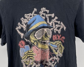 May include: Black graphic t-shirt featuring a skeleton wearing a blue hat and sunglasses, smoking a cigarette. The design includes the text "Marc Ecko" and "N.Y.C.", with a playing card and dollar sign.