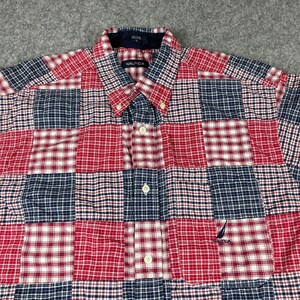 May include: A long-sleeve button-down shirt featuring a patchwork design of red, white, and navy blue plaid patterns. The shirt includes a collar, a front button placket, and a small embroidered Nautica logo on the lower right chest area.