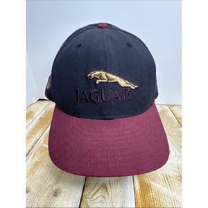 May include: A navy blue baseball cap with a maroon bill. The cap features the word "JAGUAR" embroidered in maroon, with a gold jaguar logo above it. The cap has a maroon button on top.
