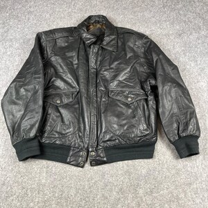 May include: A black leather bomber jacket featuring a classic collar, a front zipper closure, and two large flap pockets on the chest. The jacket includes ribbed knit cuffs and a ribbed hem, with a visible brown interior lining at the collar area.
