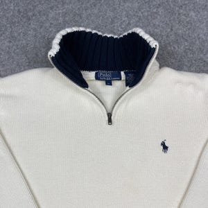 May include: Cream-colored sweater with a navy blue and white collar and a quarter-zip closure. The sweater features a small navy blue polo logo on the chest. The label reads "Polo by Ralph Lauren".