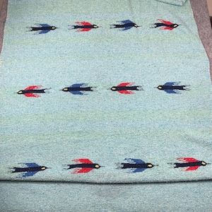 May include: A light blue woven blanket featuring a repeating pattern of stylized birds in flight. The birds are rendered in alternating solid colors of dark blue and bright red, each with a small yellow detail, arranged in three horizontal rows.
