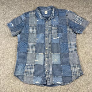 May include: A short-sleeve button-down shirt featuring a blue patchwork design with various patterns including stripes, plaids, and paisley. The shirt has a collar, a single chest pocket, and light-colored buttons down the front center placket.