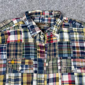 May include: A patchwork button-down shirt in a variety of plaid patterns. The shirt features a collared neckline, two chest pockets, and button closures. The color palette includes shades of blue, green, yellow, red, and brown.