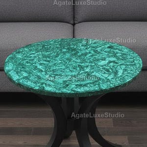 Op de afbeelding: Een ronde bijzettafel met een levendig teal oppervlak en een zwarte, gebogen poot. De tafel staat voor een grijze bank, wat een moderne en stijlvolle woonkamer creëert. De diameter is ongeveer 60 cm.