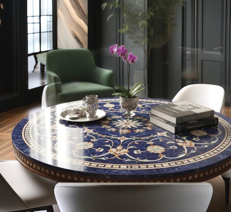 Pu&ograve; includere: Un tavolo rotondo con una superficie blu scuro con un motivo floreale color crema e oro. Il tavolo ha un bordo in legno marrone ed &egrave; circondato da sedie bianche. Un vaso di orchidee viola e libri sono sul tavolo.