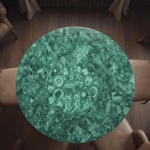 Op de afbeelding: Bovenaanzicht van een ronde tafel met een malachietblad. Het oppervlak vertoont een wervelend patroon van donkere en lichte groentinten, dat doet denken aan natuursteen. De tafel wordt omringd door vier stoelen met lichtbruine leren zittingen en houten frames.