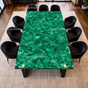 Op de afbeelding: Een rechthoekige, smaragdgroene eettafel met een mozaïekachtig oppervlak, omringd door acht zwarte fluwelen stoelen. De tafelpoten zijn zwart en de stoelen hebben goudkleurige poten. De tafel staat op een neutraal gekleurd tapijt.