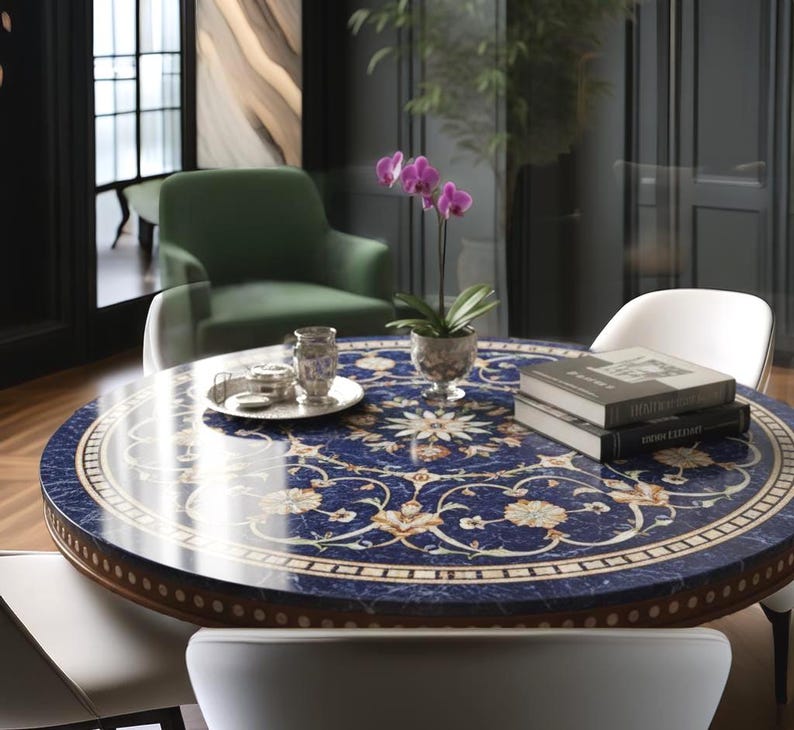 Pu&ograve; includere: Un tavolo rotondo con una superficie blu scuro con un motivo floreale bianco e oro. Il tavolo ha un bordo in legno ed &egrave; circondato da sedie bianche. Un vaso di orchidee viola e libri sono sul tavolo.