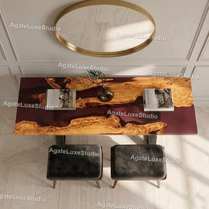 Puede incluir: Vista aérea de una mesa de comedor moderna con un centro de resina burdeos y bordes de madera natural. Dos taburetes tapizados de color gris oscuro están colocados debajo. Un espejo con marco dorado cuelga encima.