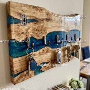 Peut inclure: Un porte-clés en bois avec un design de bord brut, avec une rivière de résine bleue. Le porte-clés comporte plusieurs crochets en métal pour accrocher les clés. Le bois est de couleur brun clair naturel.