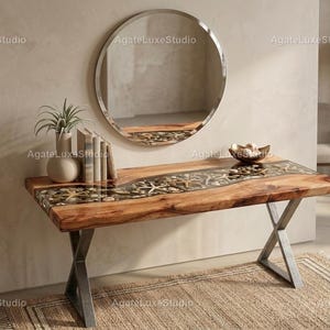 Può includere: Una consolle in legno con un design a fiume in resina trasparente, con stelle marine e conchiglie incorporate. Il tavolo ha una finitura a venature naturali e poggia su una base in metallo. Uno specchio rotondo è appeso sopra, con una pianta, libri e una ciotola decorativa.
