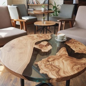 Puede incluir: Mesa de centro redonda de madera con un diseño único. La mesa presenta un patrón de vetas de madera marrón claro con detalles de resina oscura. Una taza de café está sobre la mesa. La mesa está sostenida por patas de metal negro.