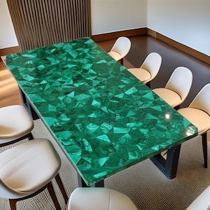 Op de afbeelding: Een rechthoekige eettafel met een levendig groen malachietblad. De tafel wordt ondersteund door een zwarte basis en is omringd door witte stoelen. Het tafelblad heeft een geometrisch patroon.